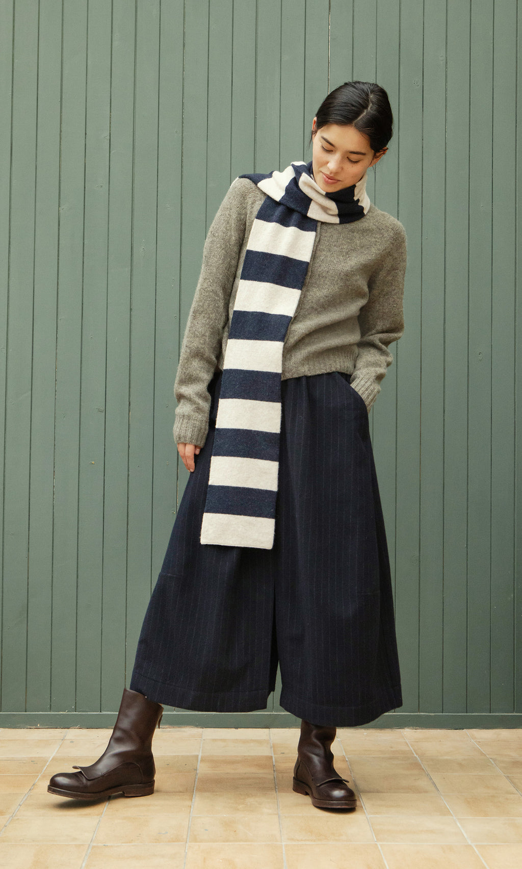 Plümo  Person in a Rugby Stripe Scarf, gray sweater, black wide-leg pants, and brown boots stands by a green paneled wall.
