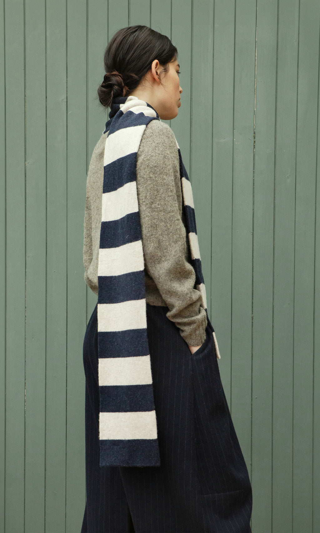 Plümo  Person in side profile wears a Rugby Stripe Scarf, gray sweater, and dark pants before a green paneled wall.