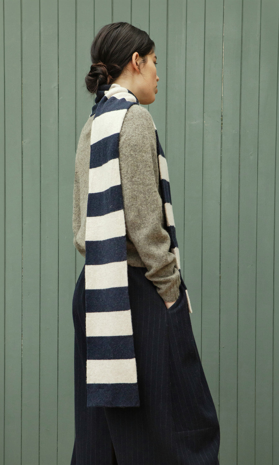 Plümo  Person in side profile wears a Rugby Stripe Scarf, gray sweater, and dark pants before a green paneled wall.