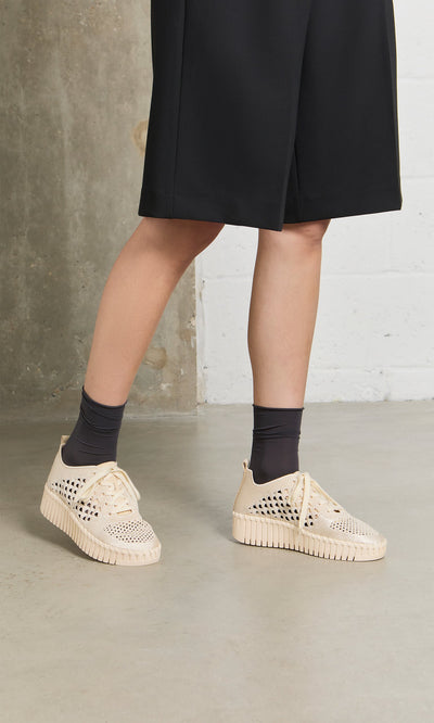 Plümo- Person wears Runa Trainers with breathable embroidered upper, black socks, black skirt, standing indoors on concrete.