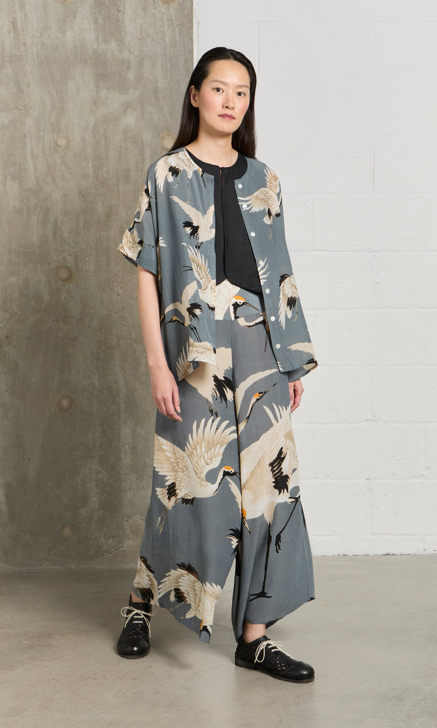 Plümo- A person wears the Slate Stork Blouse, featuring a stork motif in slate grey, standing indoors on a concrete floor.