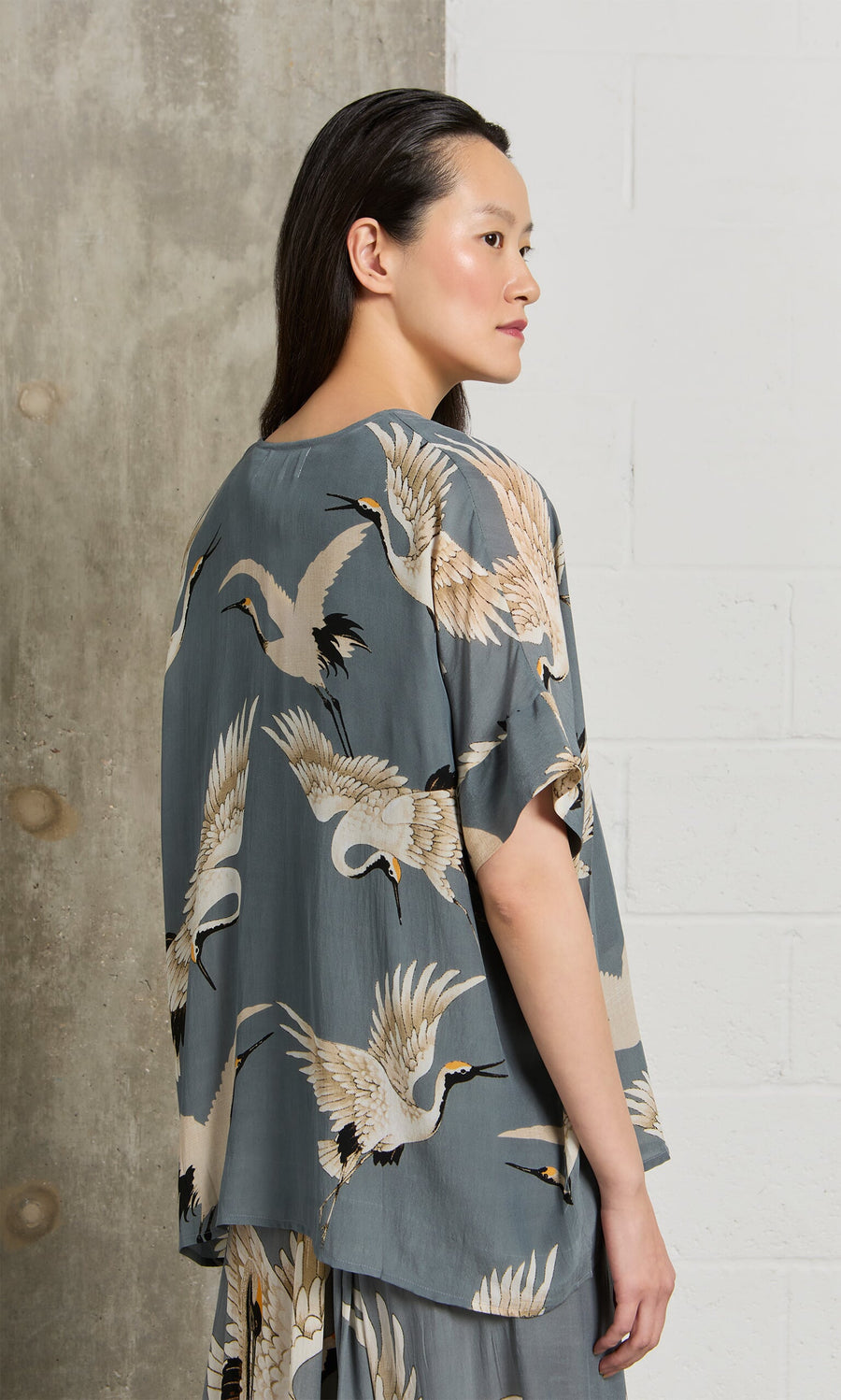Plümo- A woman stands sideways wearing the Slate Stork Blouse against concrete and white wall backgrounds.