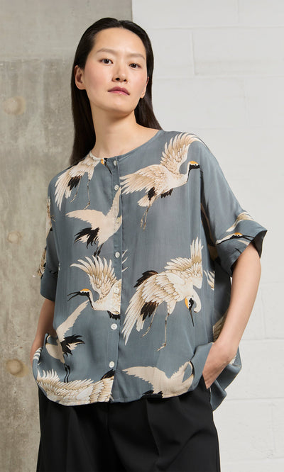 Plümo- Woman in the Slate Stork Blouse stands with hands in pockets against a concrete and white wall.