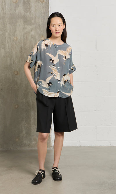 Plümo- Woman indoors wearing the Slate Stork Blouse, black knee-length shorts, and black shoes.
