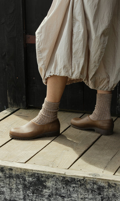 Plümo  Person wearing brown socks and Hemp Clogs stands on a wooden deck, dressed in a beige skirt near a dark wall.