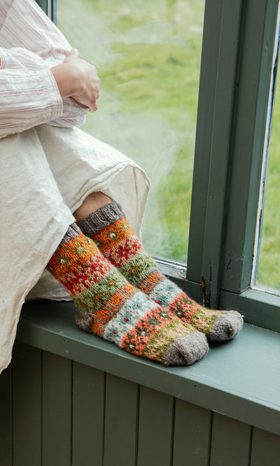 Plümo  Person sitting by a window in a cream skirt and Sofa Socks, hands on knees, savoring the moment.