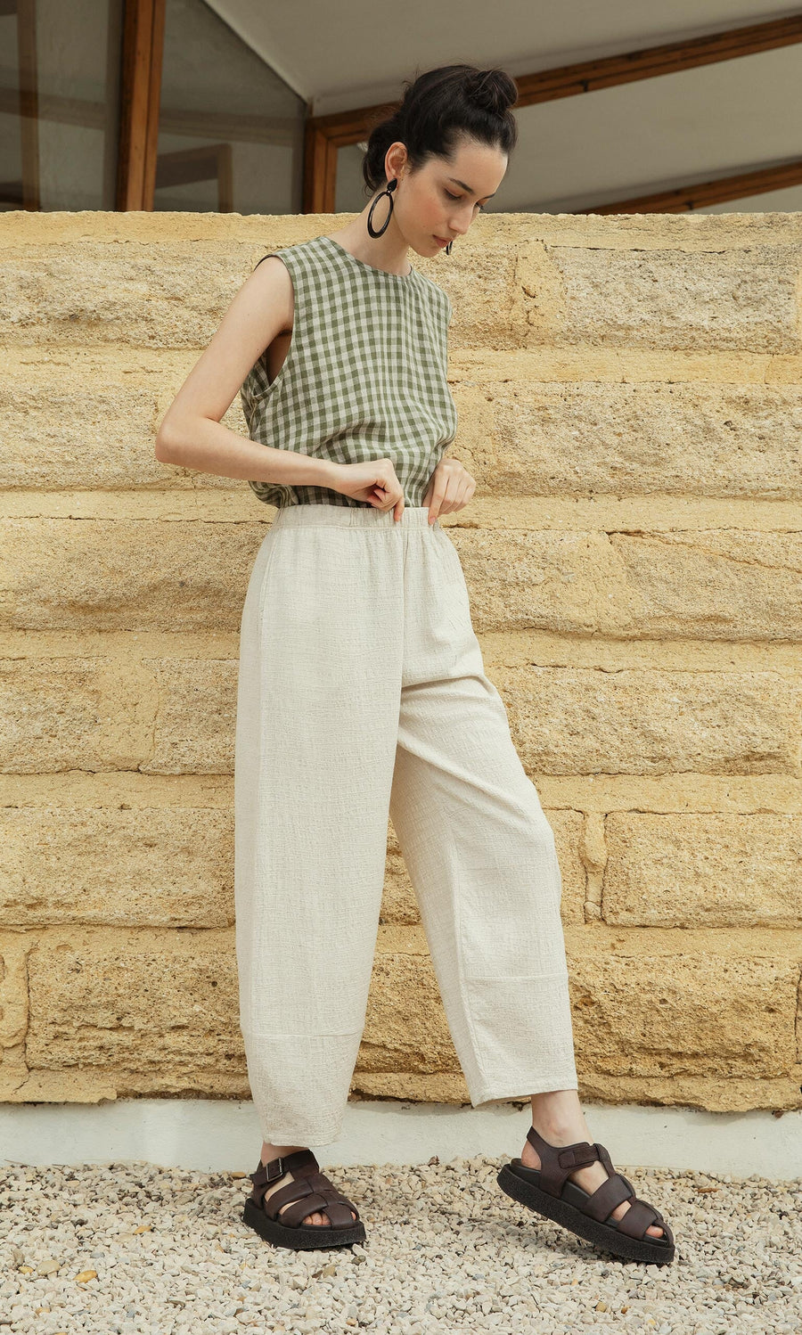 Plümo- Woman in cream Soren Pants, a sleeveless green checkered top, and black sandals by a textured stone wall.