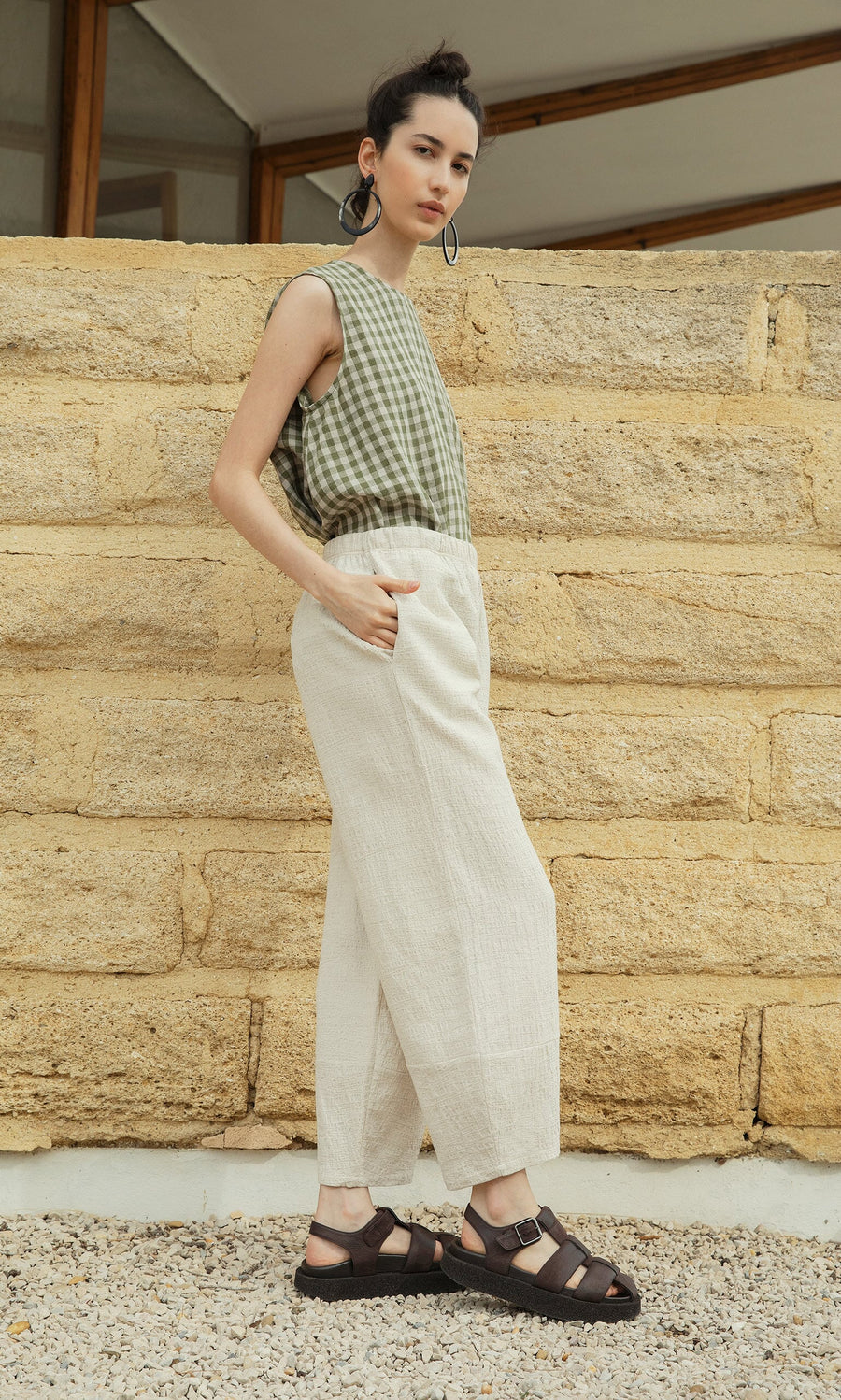 Plümo- Woman in green checkered sleeveless top, Soren Pants, and sandals stands by a tan stone wall, hands in pockets.
