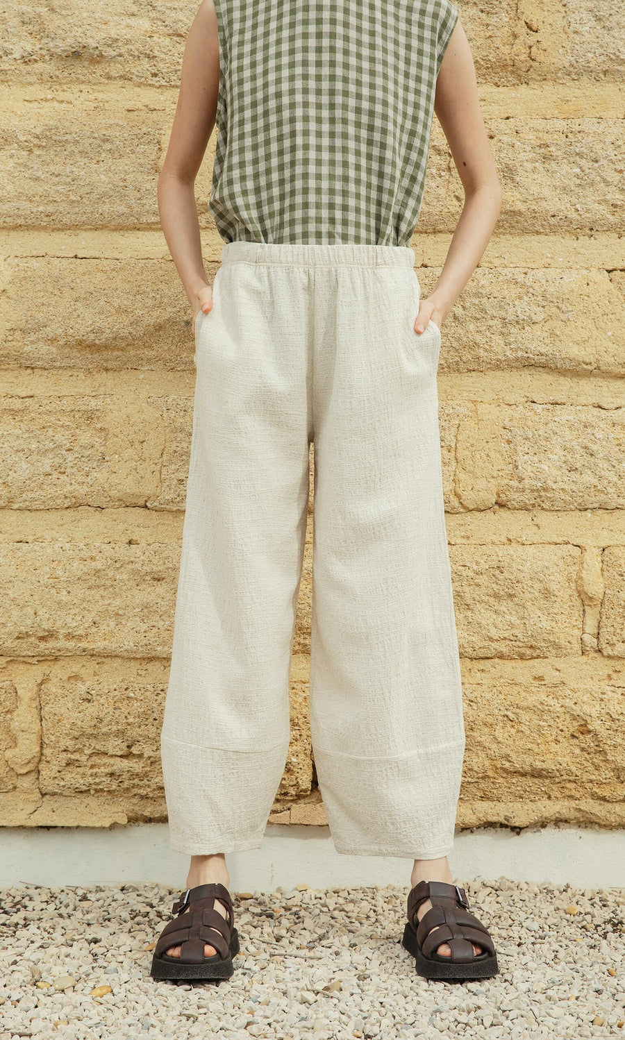 Plümo- Person wearing a green checkered sleeveless top, Soren Pants in cream, and black sandals against a stone wall.