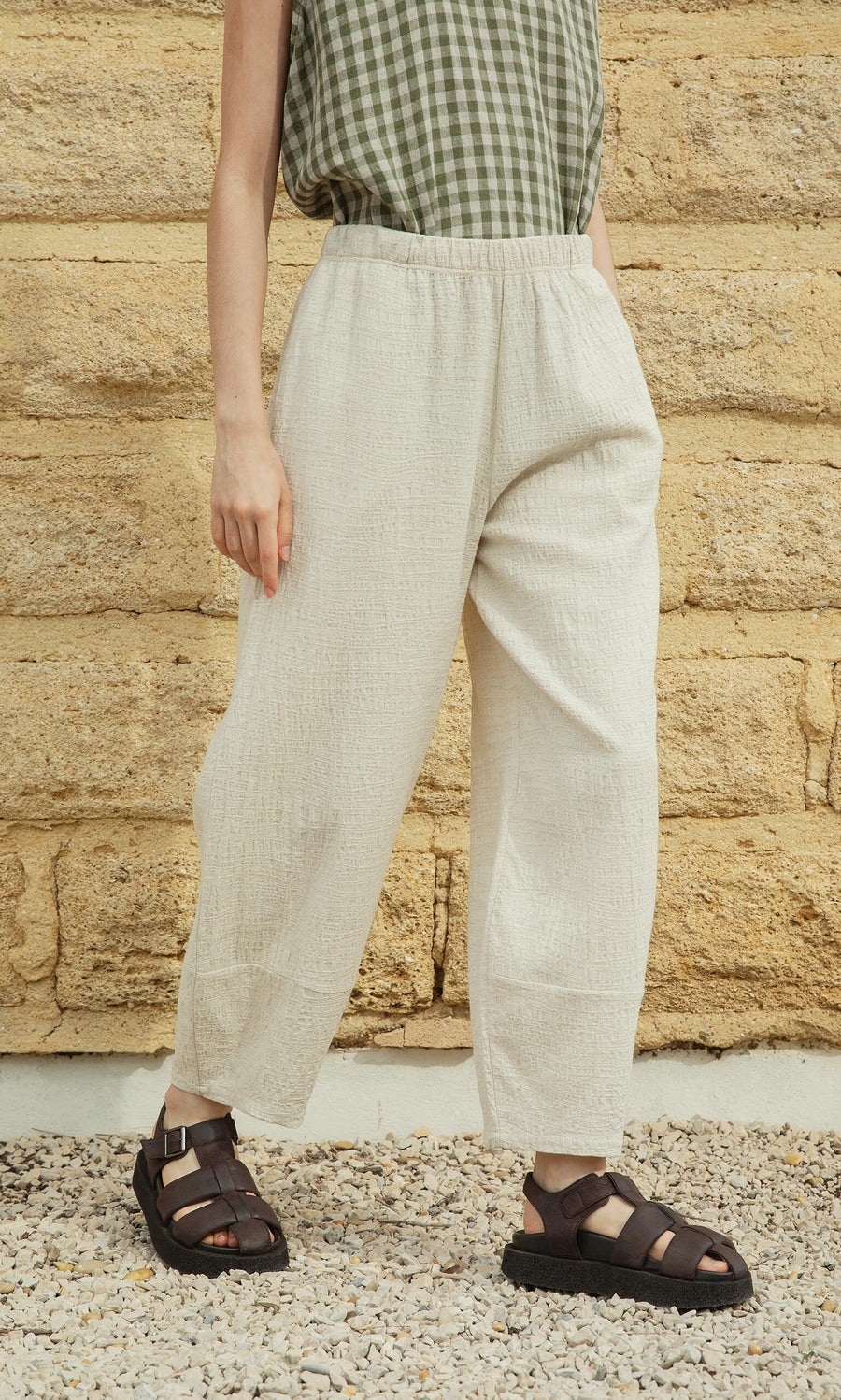Plümo- Person in a green checkered top, Soren Pants in cream, and black sandals stands on gravel by a tan stone wall.