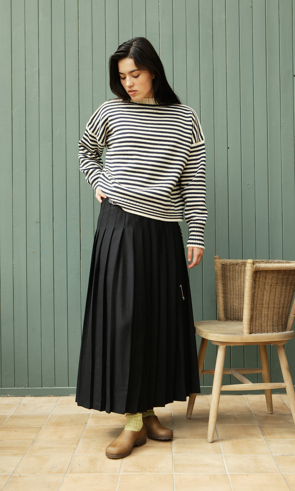 Plümo  Woman in Carnac Jumper and black pleated skirt stands by a chair, looking down, green wall behind.