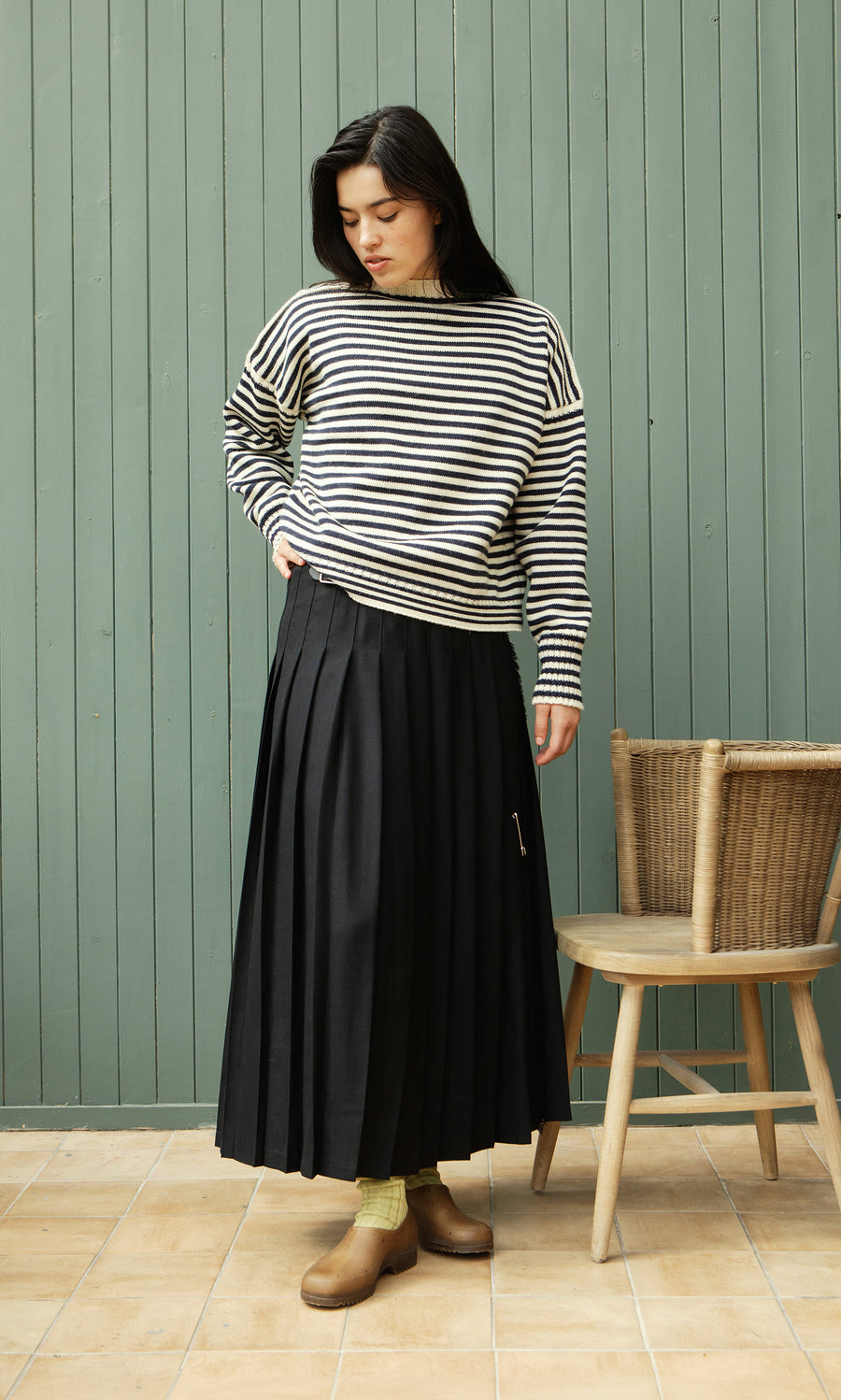 Plümo  Woman in Carnac Jumper and black pleated skirt stands by a chair, looking down, green wall behind.