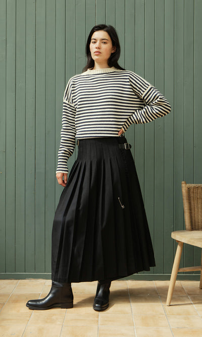 Plümo  Woman in a striped sweater, Storm Kilt, and boots stands by a green paneled wall and wooden chair.