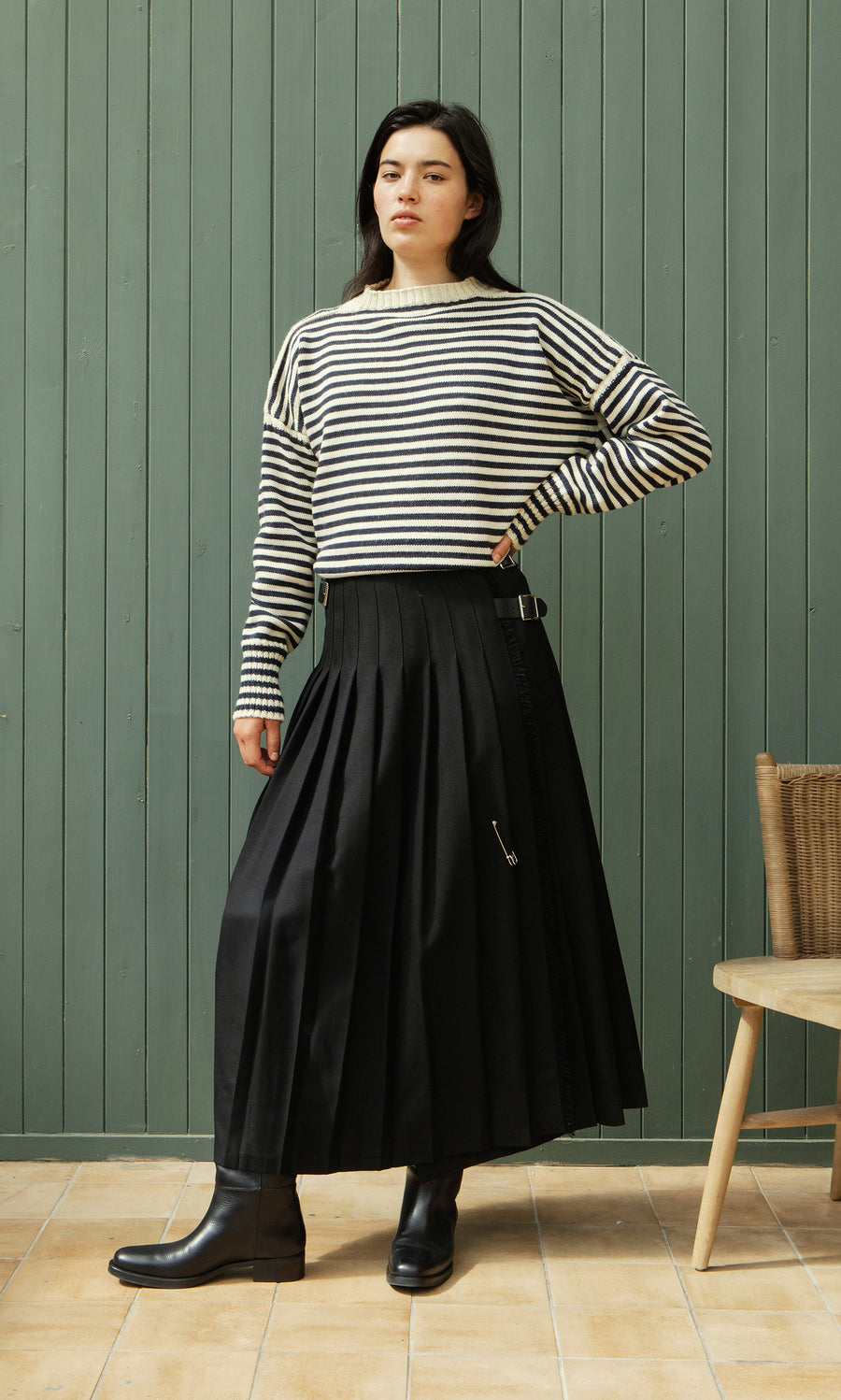 Plümo  Woman in a striped sweater, Storm Kilt, and boots stands by a green paneled wall and wooden chair.