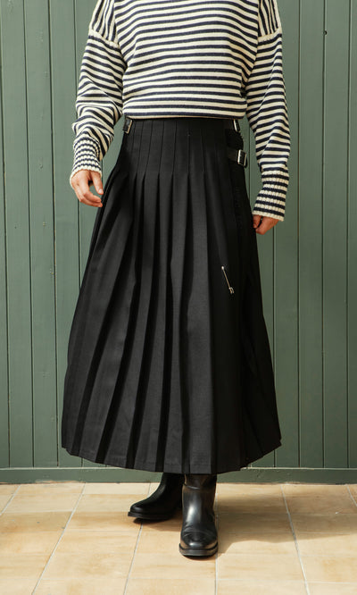 Plümo  Person in a black Storm Kilt, striped sweater, and boots stands before a green wooden wall.