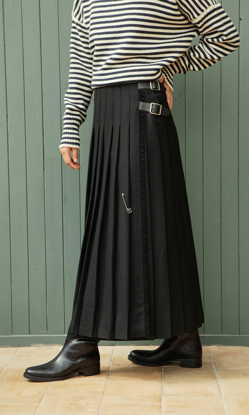 Plümo  Person wearing a striped top, Storm Kilt maxi skirt, and black boots stands by a green wooden wall.