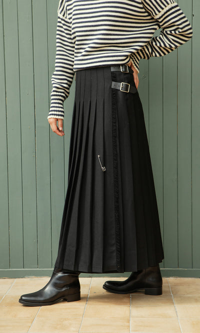 Plümo  Person wearing a striped top, Storm Kilt maxi skirt, and black boots stands by a green wooden wall.