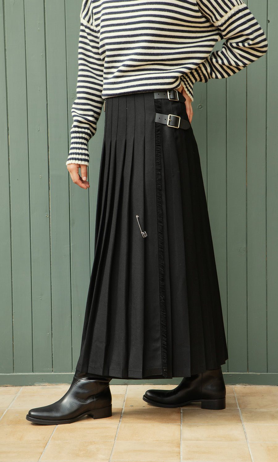 Plümo  Person wearing a striped top, Storm Kilt maxi skirt, and black boots stands by a green wooden wall.