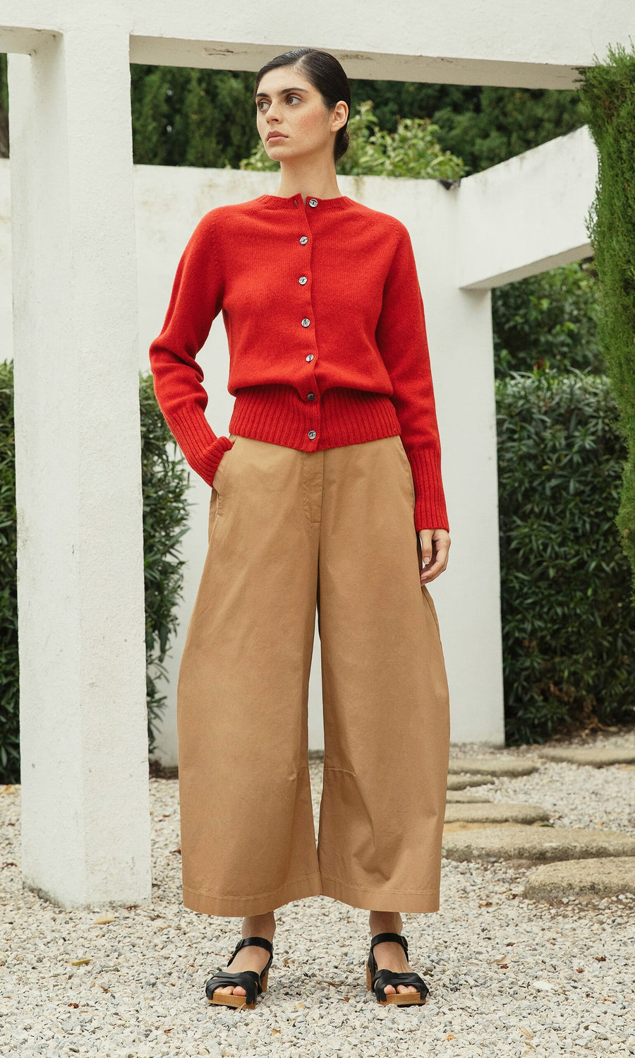 Plümo- Woman in a red cardigan, Karate Pants, and black sandals stands outdoors near white pillars and greenery.
