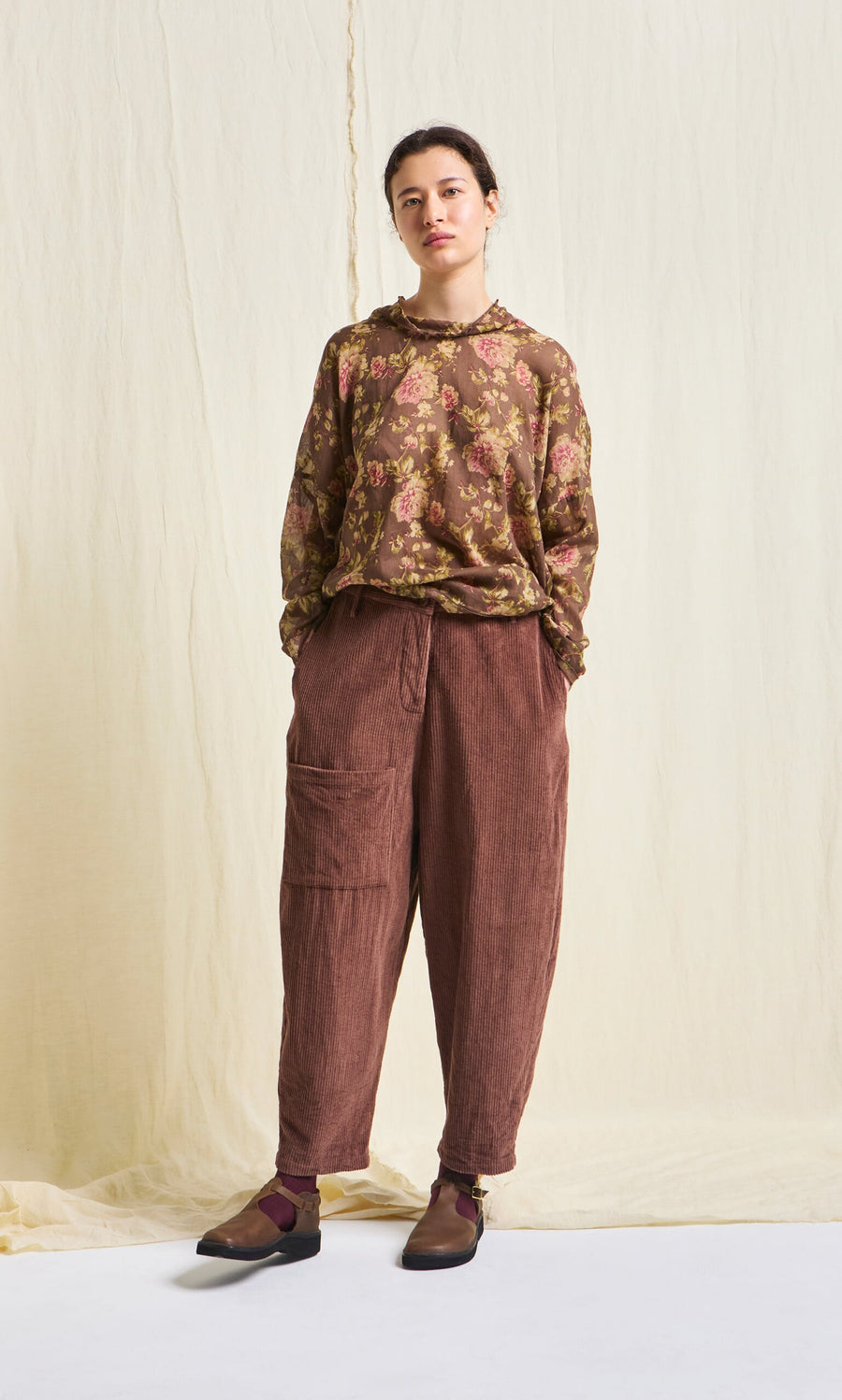 Plümo- Woman wearing floral blouse, Lumber Pants, brown shoes, and burgundy socks against a cream backdrop.