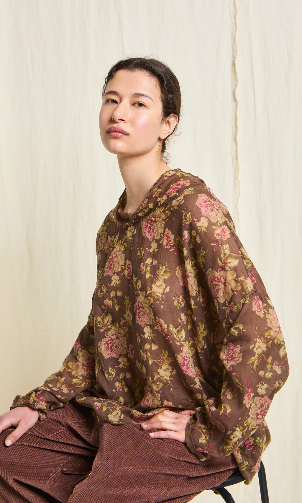 Plümo- Woman sits on a chair in the Sylvie Shirt, a lightweight cotton floral print top, against a light background.