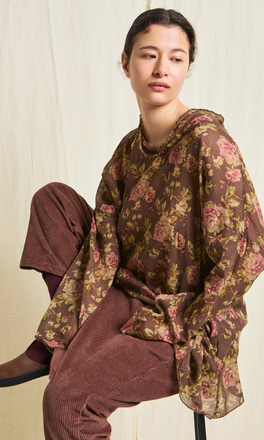 Plümo- Woman wearing Lumber Pants and a brown floral blouse, seated on a stool against a light background.