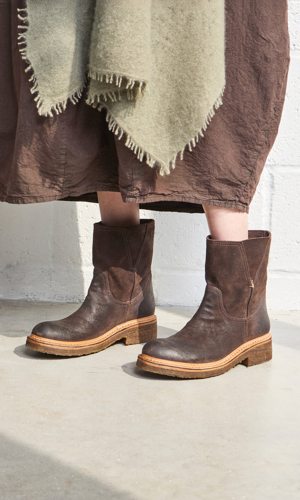 Plümo- Person wearing Tait Boots, a brown skirt, and a green fringed shawl, standing on a concrete floor.