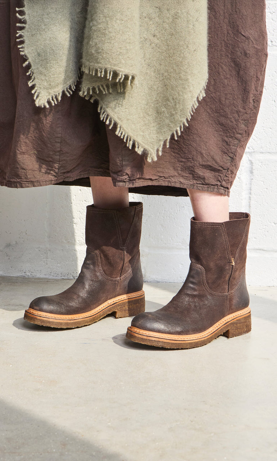 Plümo- Person wearing Tait Boots, a brown skirt, and a green fringed shawl, standing on a concrete floor.