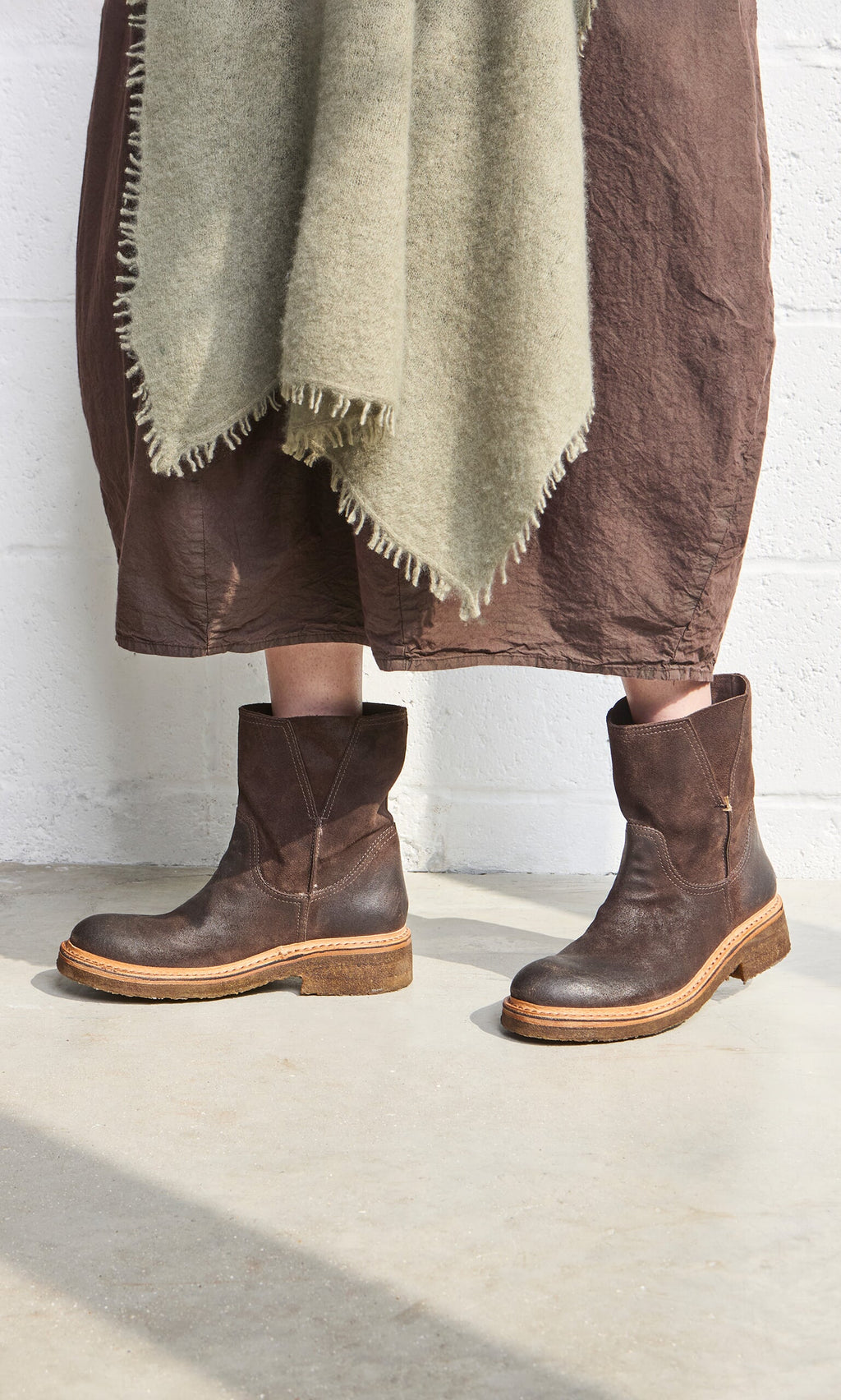 Plümo- Person wearing Tait Boots, a brown skirt, and a fringed greenish shawl, standing indoors on a concrete floor.
