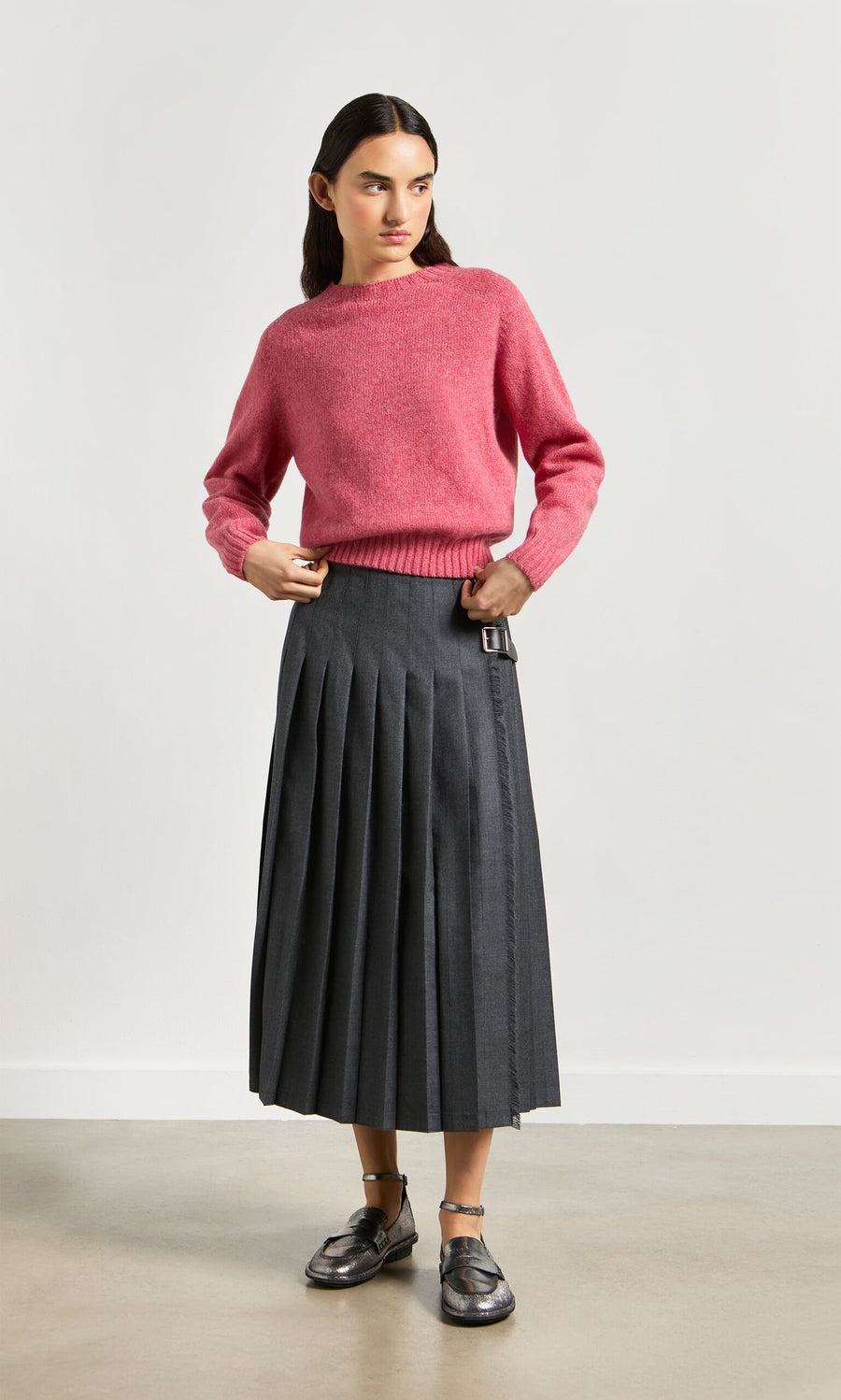 Plümo- Woman wears the Wicklow Crewneck in Tea Rose, gray pleated midi skirt, and metallic sandals against a light background.