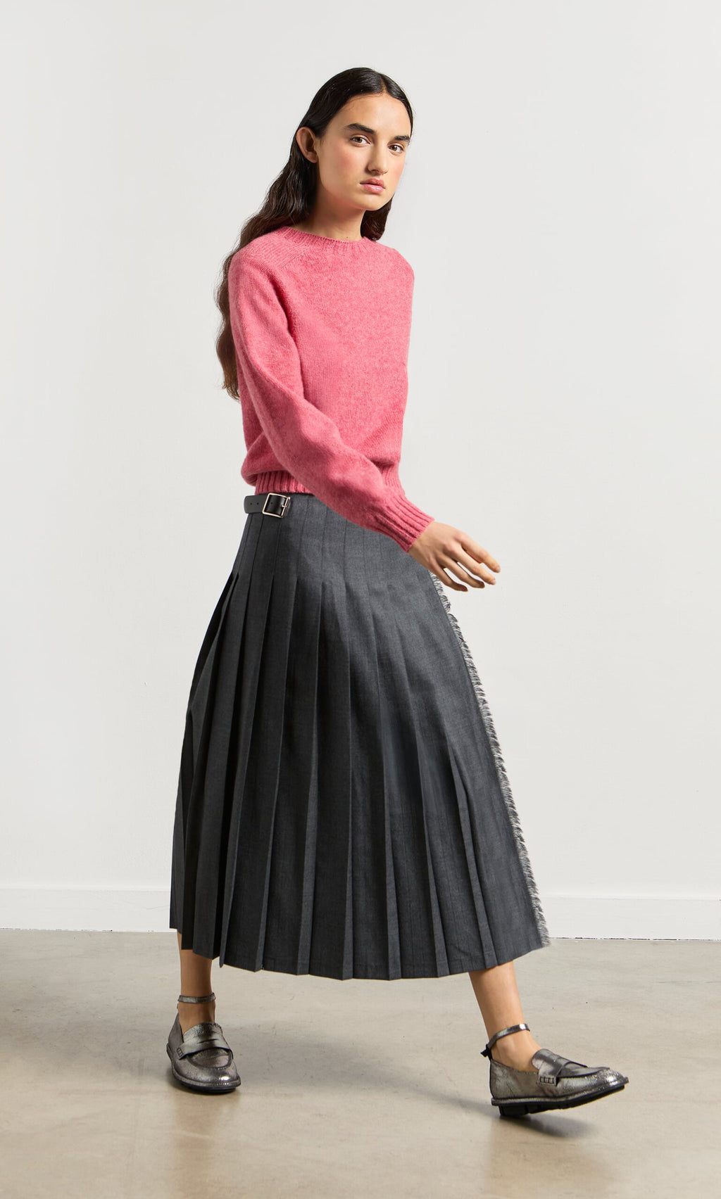 Plümo- Woman in a pink sweater, Boyd Kilt in grey worsted wool made in Ireland, and metallic shoes walks by a white wall.