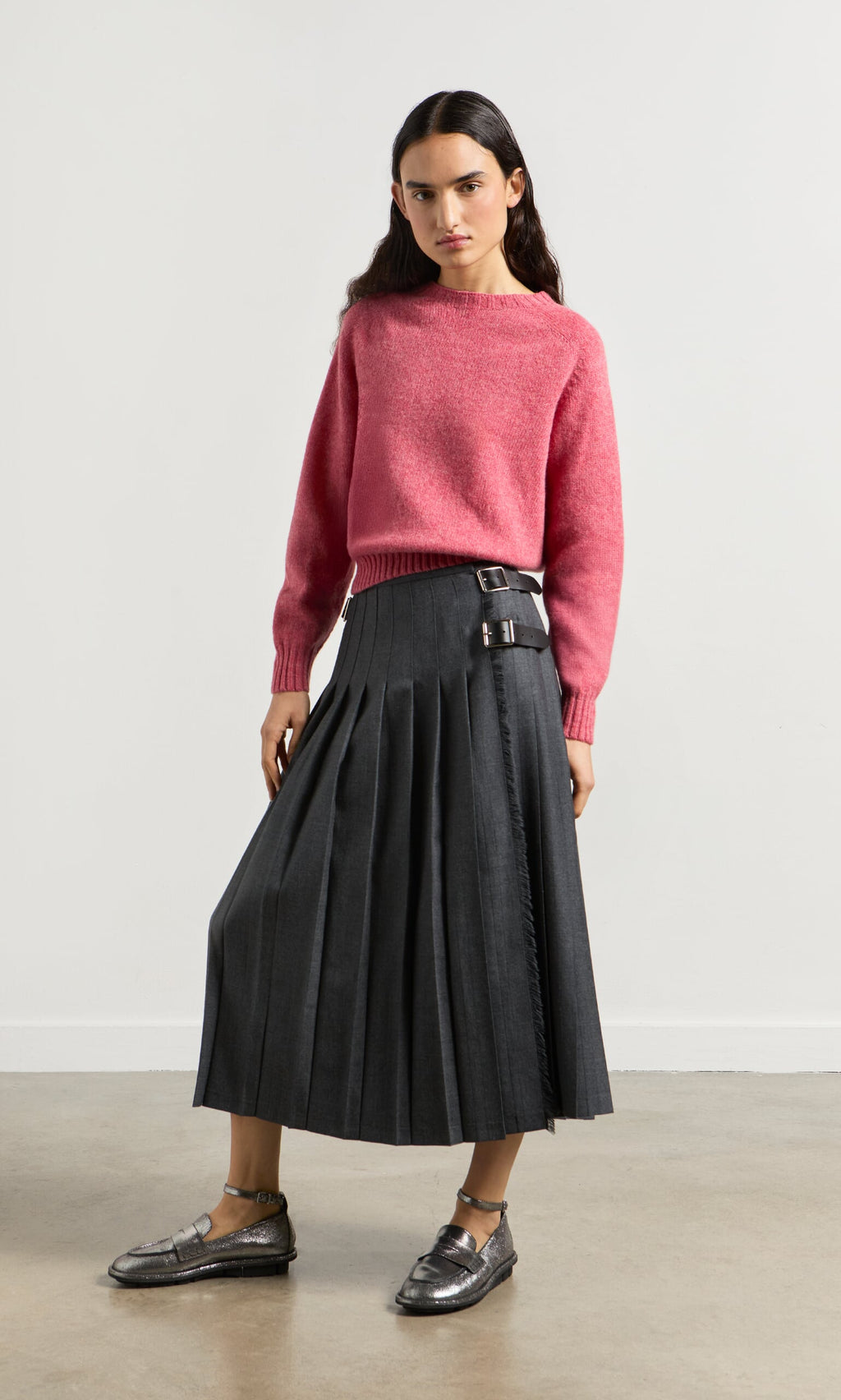Plümo- Woman in a pink sweater and Boyd Kilt, wearing metallic loafers, stands against a plain light wall.