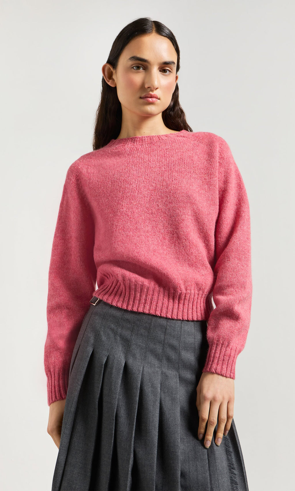 Plümo- Person in Wicklow Crewneck - Tea Rose sweater and gray pleated skirt, standing against a plain light background.