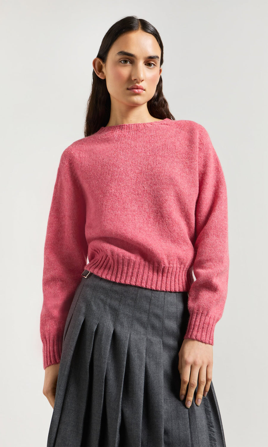 Plümo- Person in Wicklow Crewneck - Tea Rose sweater and gray pleated skirt, standing against a plain light background.