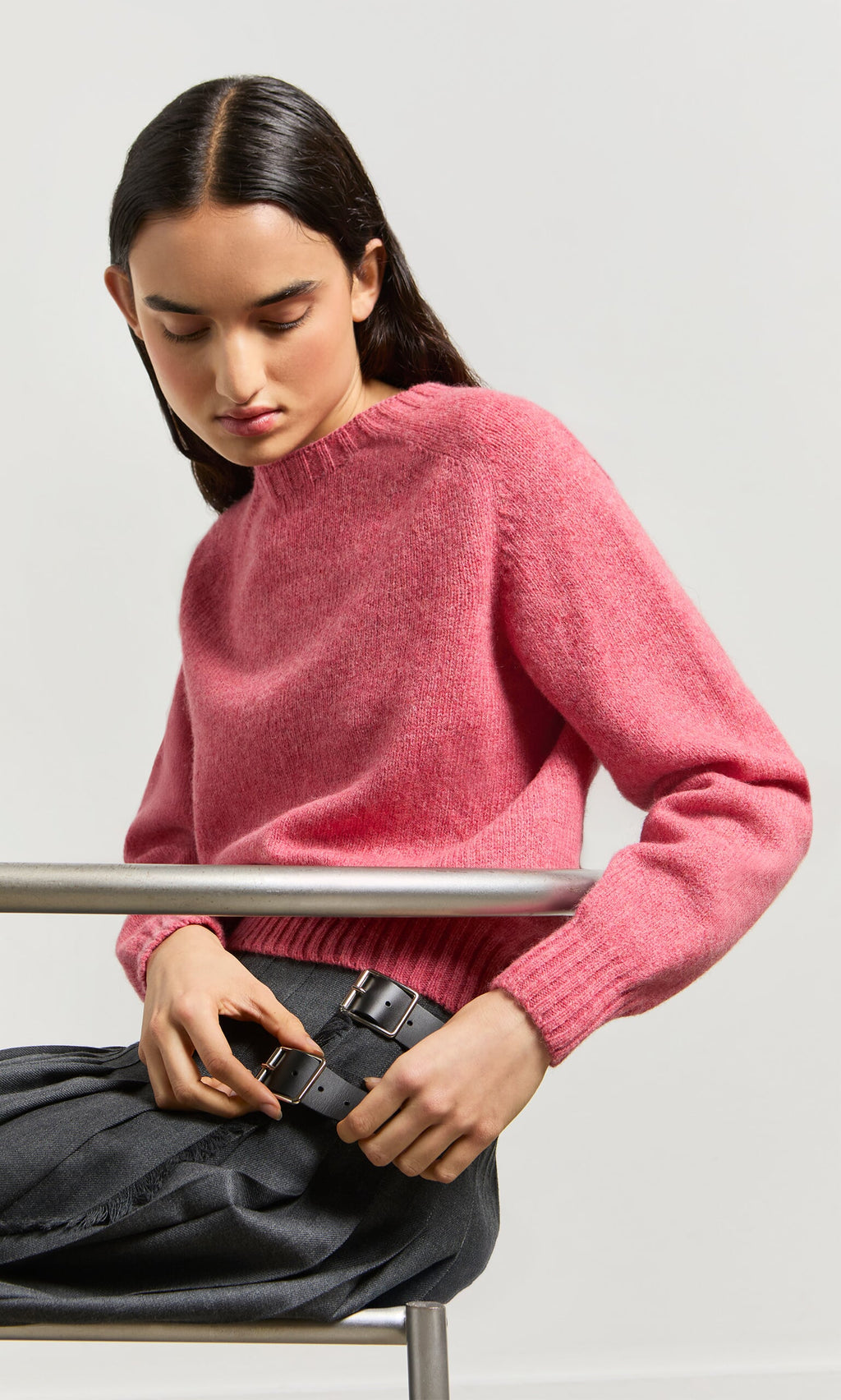 Plümo- A person in a Tea Rose Wicklow Crewneck sweater and gray pleated skirt sits, looking down, holding a large black belt.