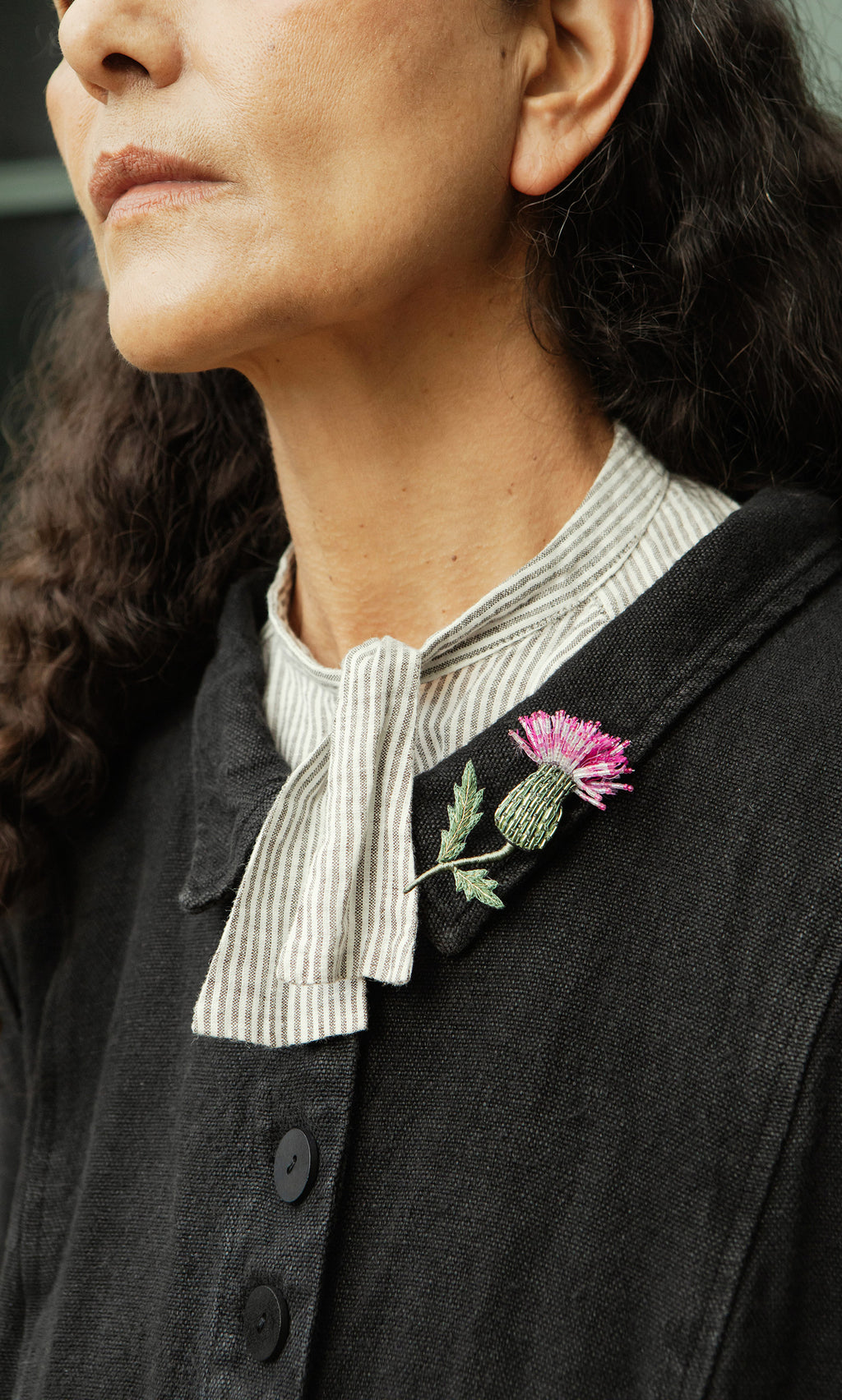 Plümo  Woman in a dark coat with a striped shirt, wearing a Thistle Brooch on her collar.