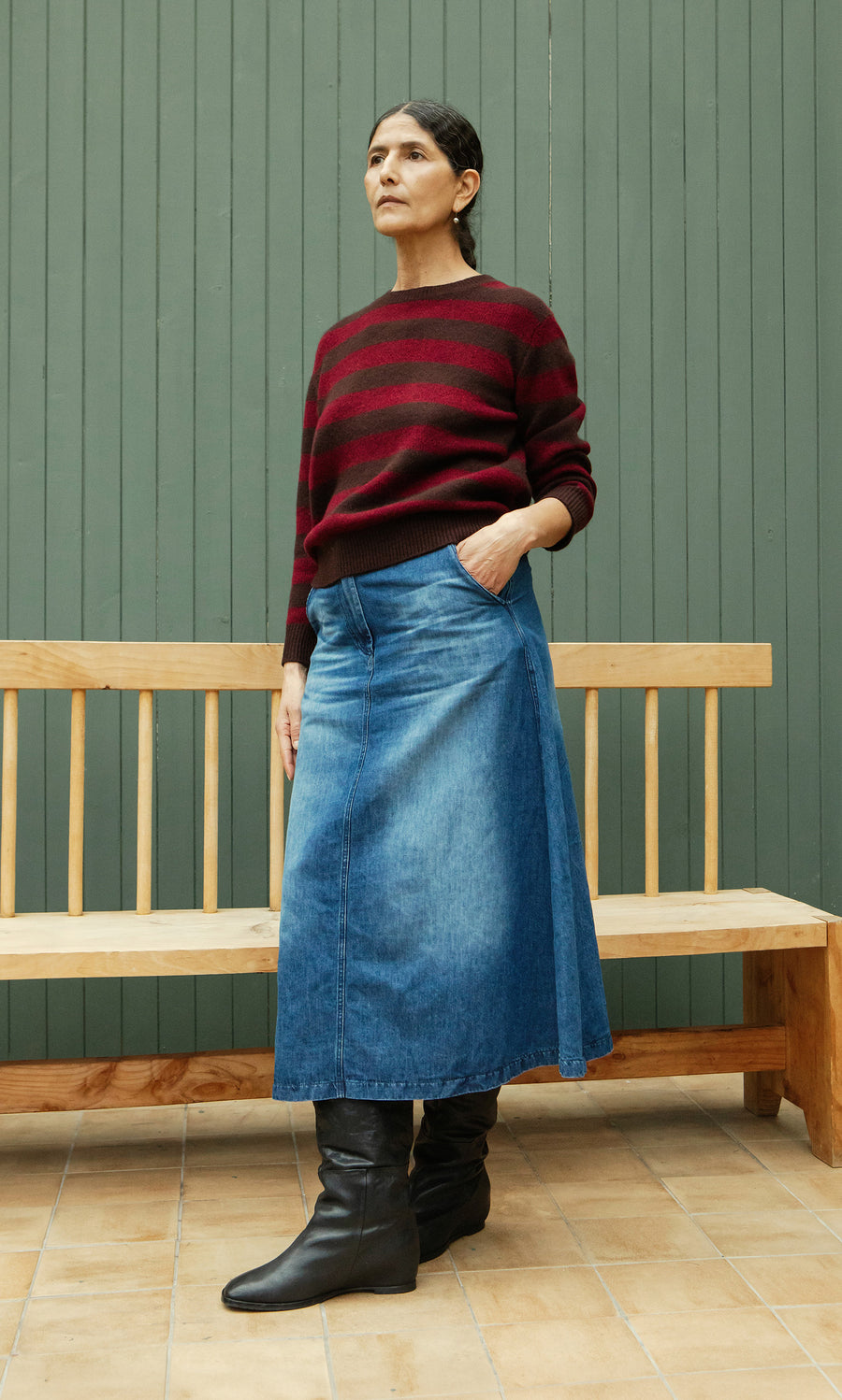 Plümo  Woman in a Tilburg Stripe Jumper, denim skirt, and boots stands by a wooden bench against a green wall.
