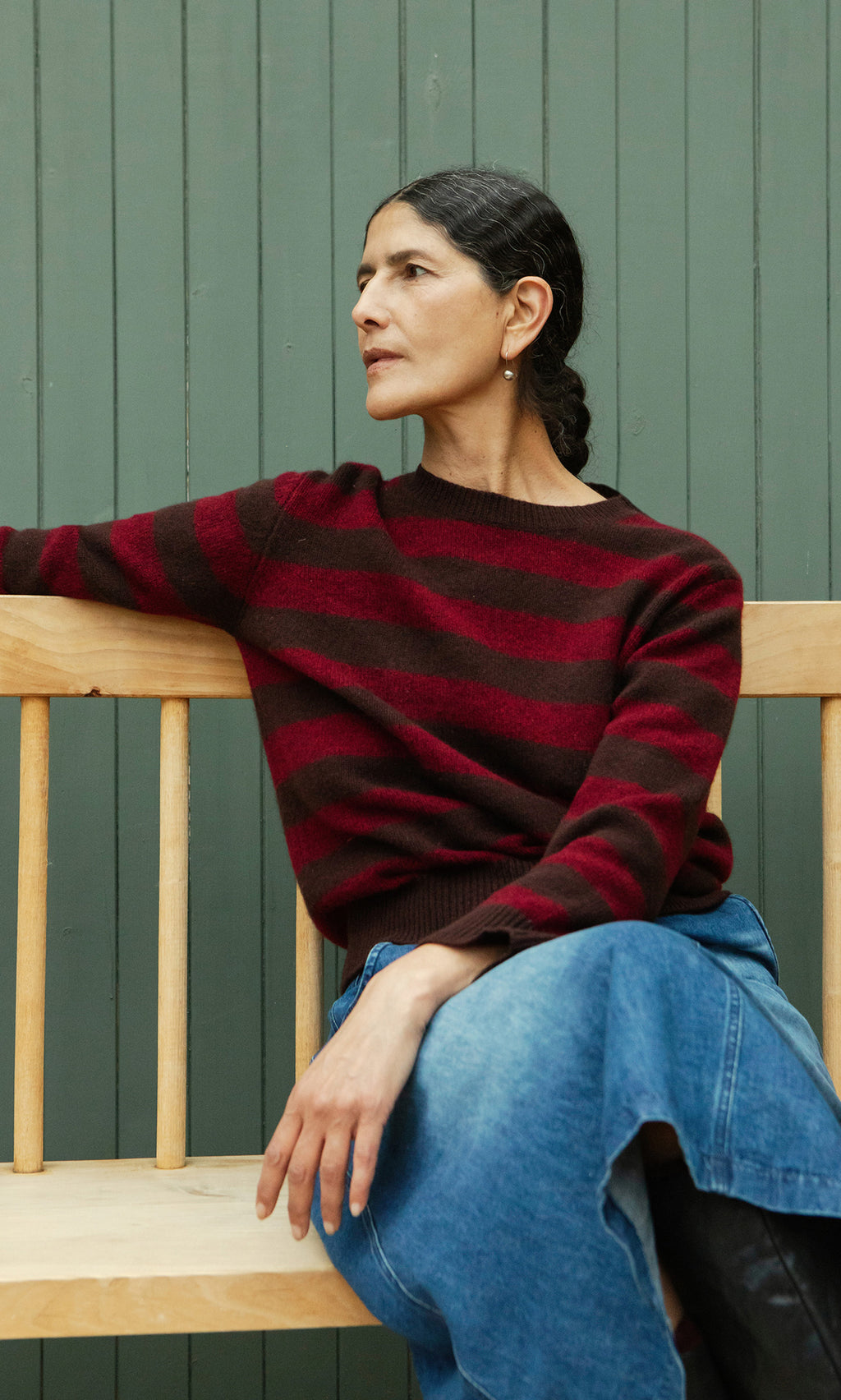Plümo  Woman in the Tilburg Stripe Jumper sits on a wooden bench, looking aside, against a green paneled background.
