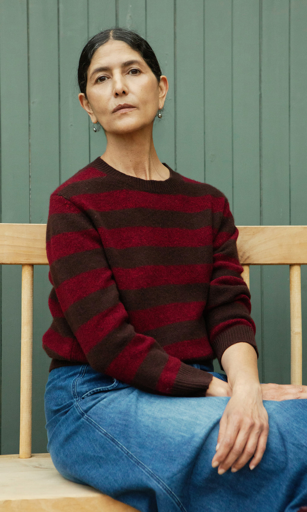 Plümo  A person with dark hair in the Tilburg Stripe Jumper and jeans sits on a wooden bench by a green paneled wall.