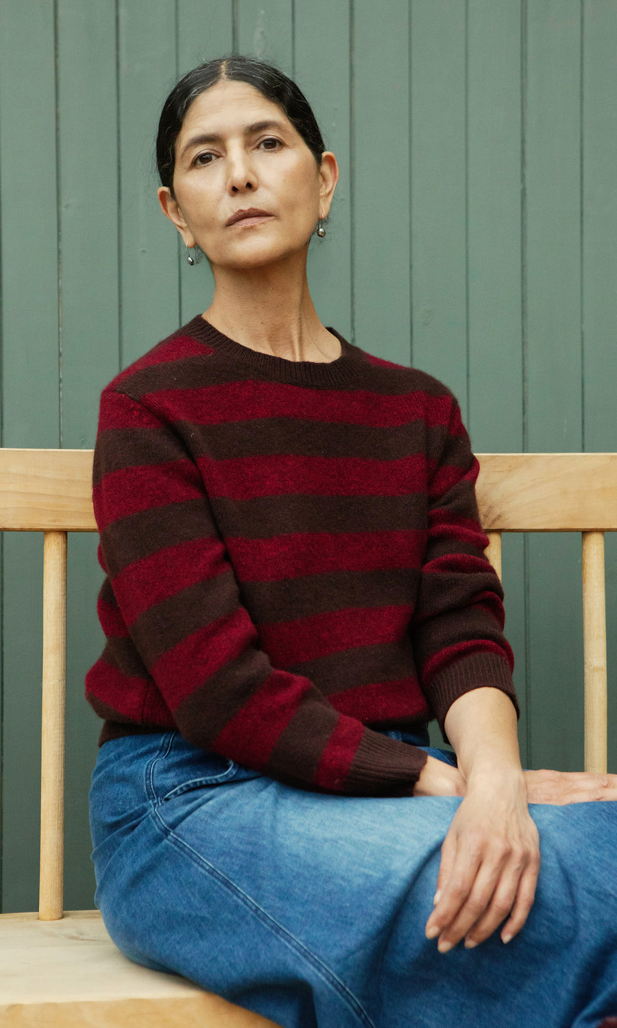 Plümo  A person with dark hair in the Tilburg Stripe Jumper and jeans sits on a wooden bench by a green paneled wall.