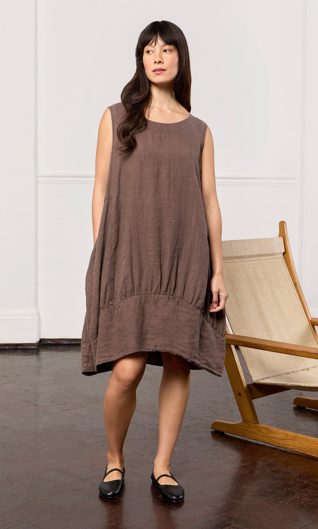 Plümo- Woman in a relaxed fit Toledo Dress and black shoes stands indoors next to a wooden lounge chair.