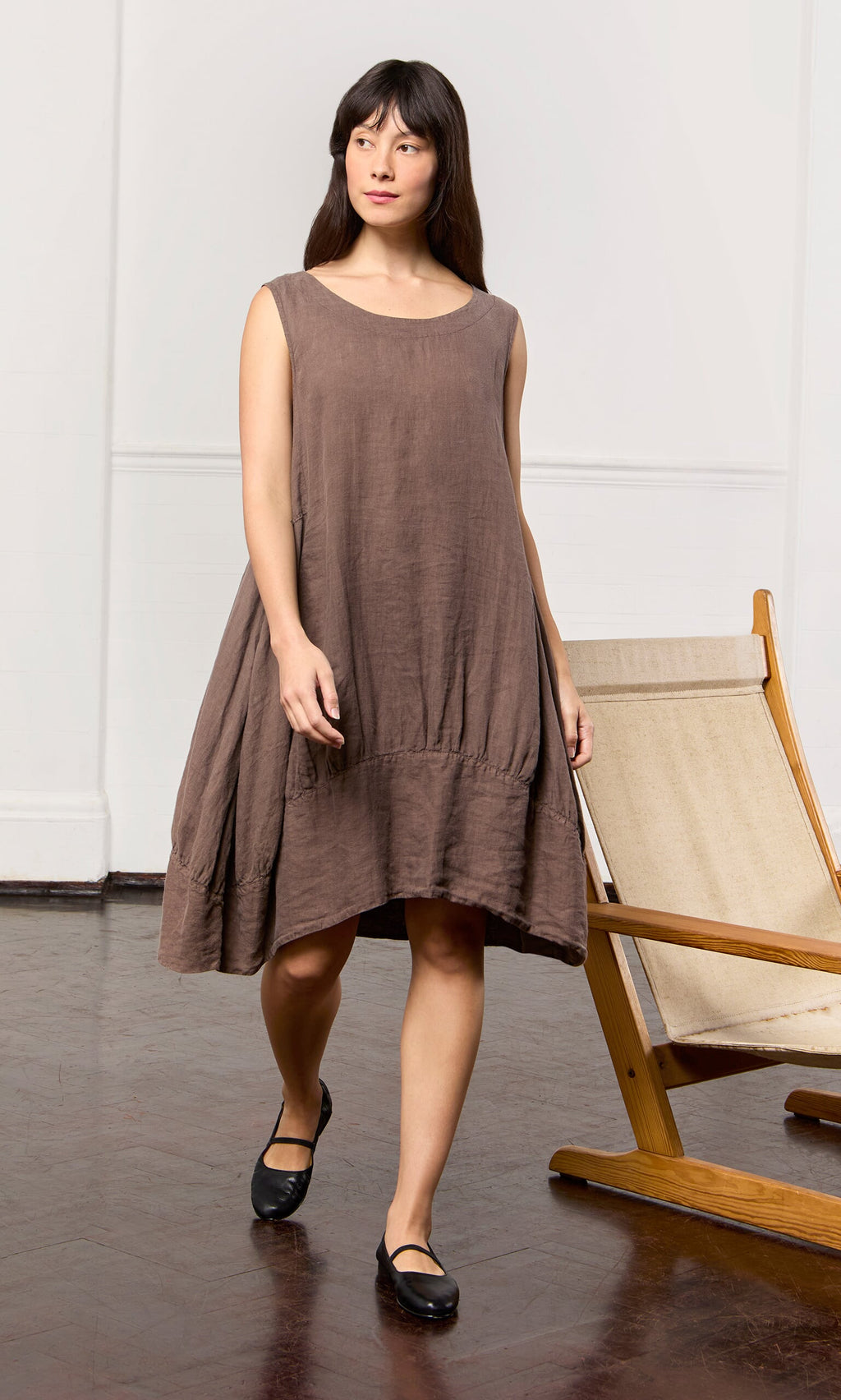 Plümo- A woman in a relaxed fit Toledo Dress stands indoors by a wooden chair, looking to the side.