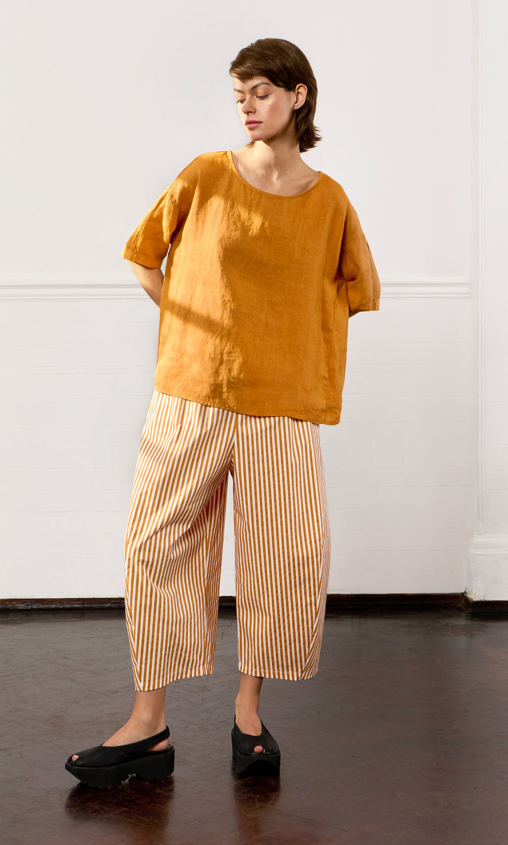 Plümo- Person wearing the Tulley Top in orange and orange-and-white striped pants, standing indoors with hands behind their back.