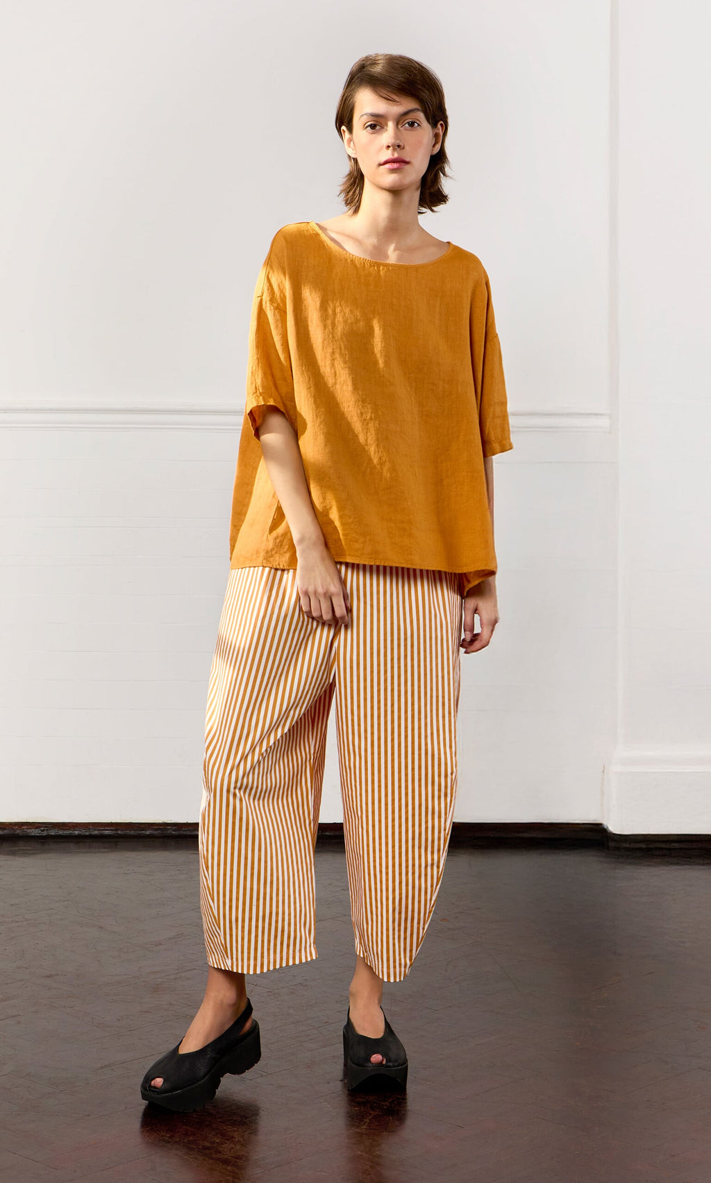 Plümo- Person wears the Tulley Top, an oversized mustard yellow boat neck top, and striped pants indoors by a white wall.