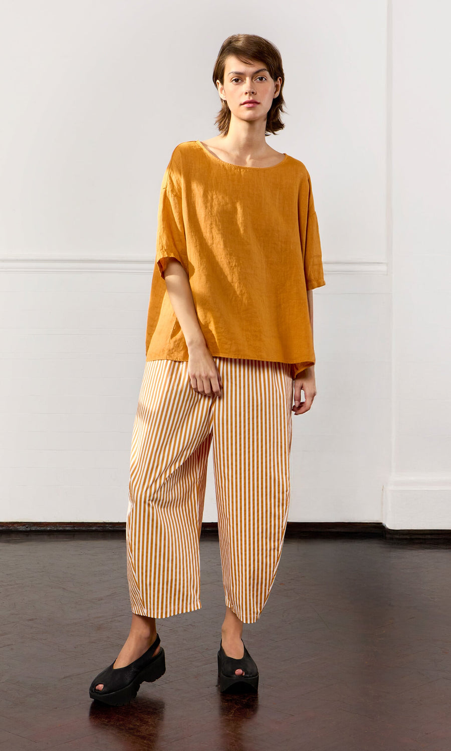 Plümo- Person wears the Tulley Top, an oversized mustard yellow boat neck top, and striped pants indoors by a white wall.