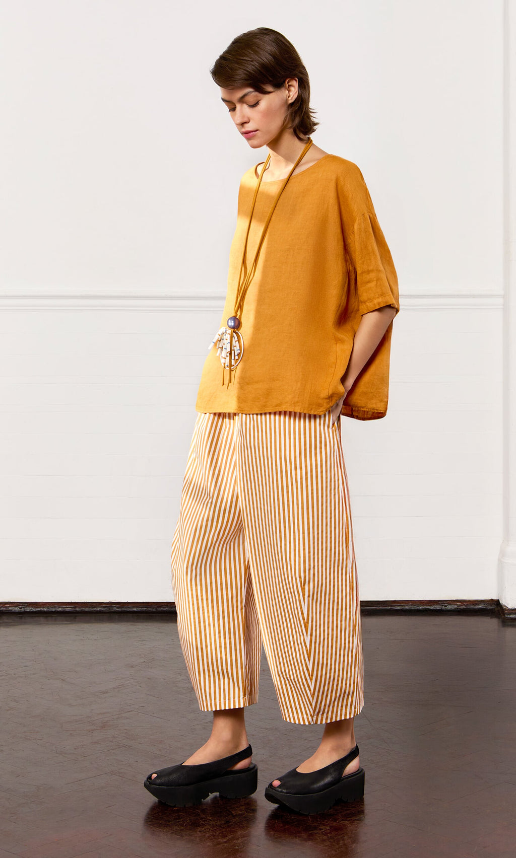 Plümo- Person in a Tulley Top, striped wide-leg pants, black sandals, and a long necklace, standing indoors.