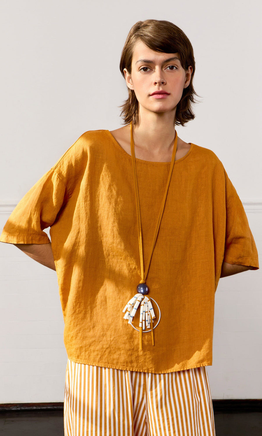Plümo- Woman in the Tulley Top—mustard yellow linen with a boat neck—paired with striped pants and a long necklace.
