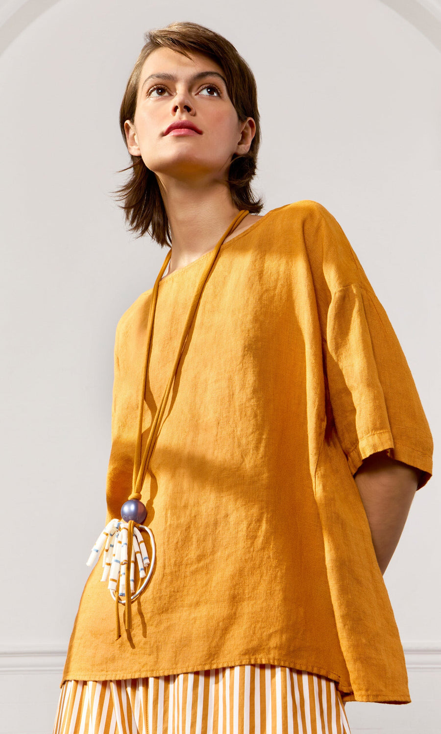 Plümo- Woman in the Tulley Top, a loose mustard yellow boat neck linen tee, with a striped skirt and statement necklace.