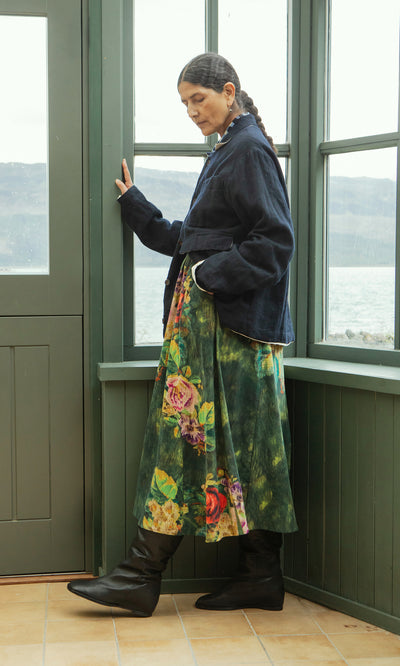 Plümo  Woman in a navy jacket and the Verity Skirt stands by green windows, looking down with one hand on the window frame.