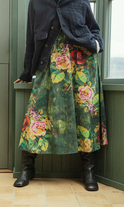 Plümo  Person in a dark jacket and Verity Skirt stands indoors by a window, wearing black boots.